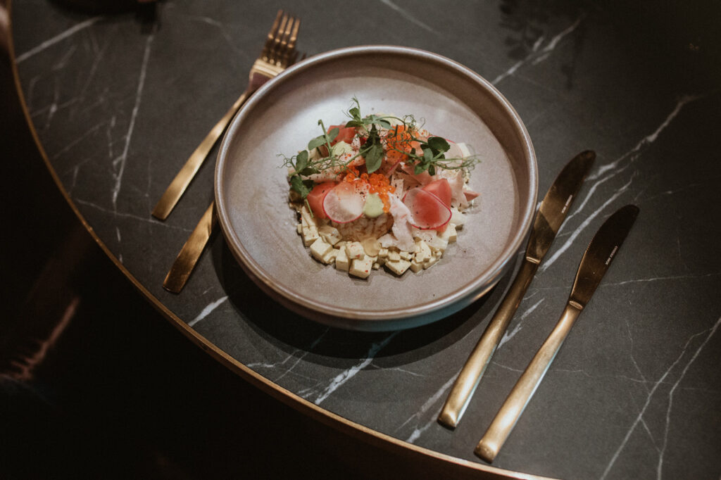 Eine stilvolle Gourmet-Vorspeise, kunstvoll auf einem grauen Teller angerichtet, bestehend aus feinen Zutaten wie Radieschen, Kaviar und frischen Kräutern. Das Gericht ist auf einem eleganten Marmortisch mit goldfarbenem Besteck präsentiert.