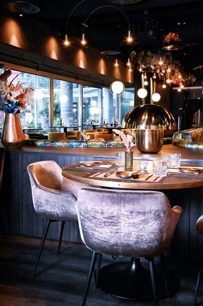 Ein stilvoll eingerichtetes Restaurant mit modernen Hängelampen, bequemen samtbezogenen Stühlen und einer festlich gedeckten Tischgruppe. Im Hintergrund sind große Fenster und eine gemütliche Atmosphäre zu sehen.