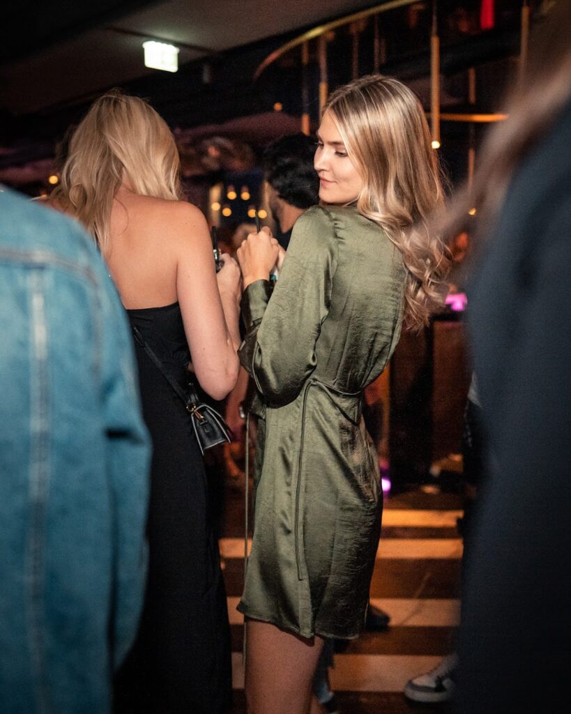 Eine junge Frau in einem eleganten, grünen Kleid genießt die Party-Nacht in einem stilvollen Club. Sie unterhält sich mit einer anderen Frau in einem schwarzen Kleid, während im Hintergrund die lebhafte Atmosphäre des Clubs zu sehen ist.